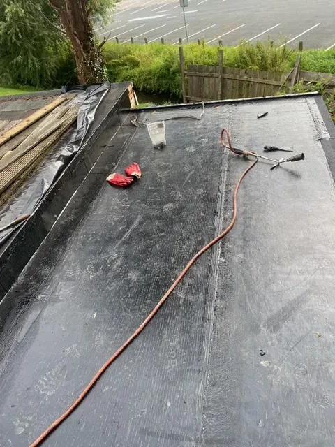 A torch-on felt flat roof part-way through, with the black underlay bonded down and the roofer's gas torch and gloves lying on it